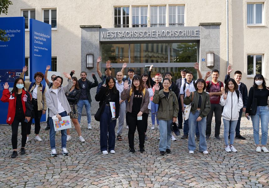 Eine Gruppe internationaler Studierende steht vor Hochschuleingang und winkt in die Kamera. 