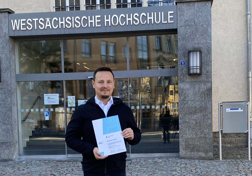 Foto: Matthias Wissel vor dem Eingangsportal Hochschule. In der Hand die Urkunde der Auszeichnung. 