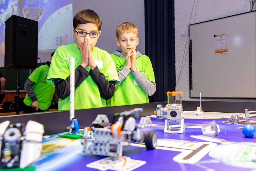 Zwei Schüler stehen mit gefalteten Händen hinter einem Lego-Spielbrett auf dem Roboter bestimmte Aufgaben erfüllen müssen. 