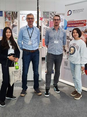 Standbetreuer Prof. Müller und Wissenschaftlicher Mitarbeiter stehen mit zwei Messebesucherinnen am Stand und blicken in die Kamera