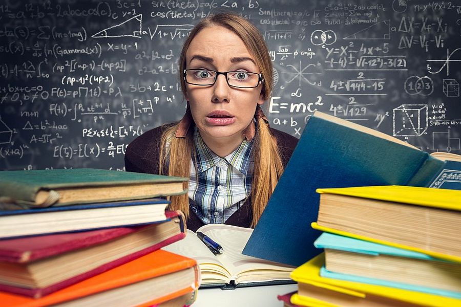 Foto: Eine Studentin sitzt verzweifelt zwischen vielen Büchern und vor einer Tafel mit Formeln.