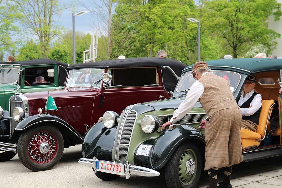 Foto: Mehrere historische PKW stehen nebeneinander. In einem sitzt ein Mann in, ein weiterer Mann steht am Auto. Beide tragen Kleidung im Stil der 1930er Jahre.