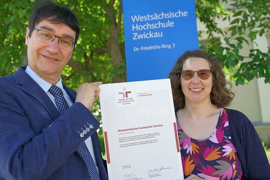 Foto: Ein Mann (links) und eine Frau (rechts) stehen vor einem Schild auf dem Westsächsische Hochschule Zwickau steht und halten ein Zertifikat hoch.