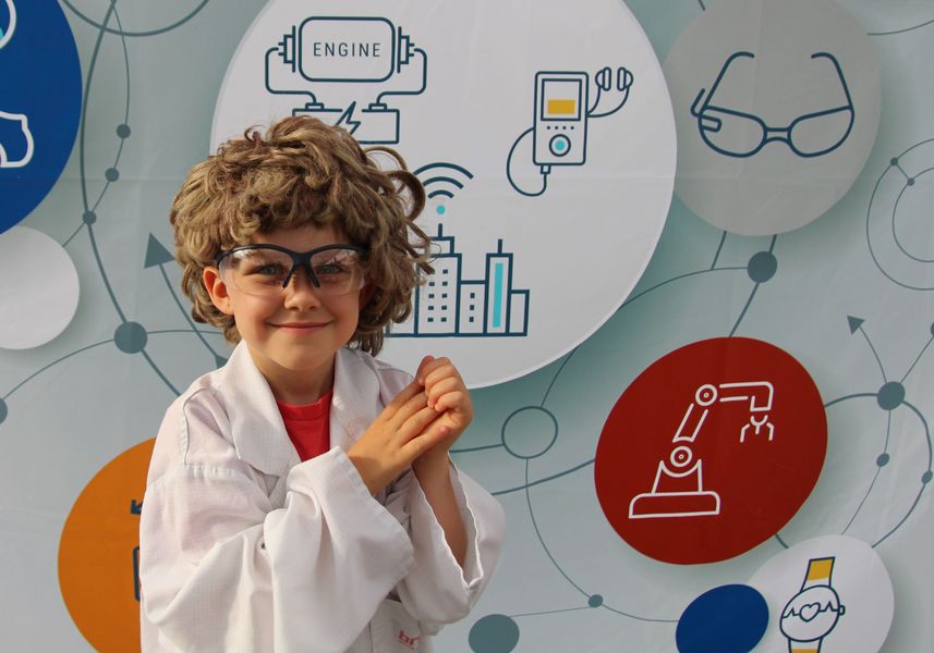 Foto: Ein Kind mit Laborbrille, Perücke und Laborkittel lächelt in die Kamera.