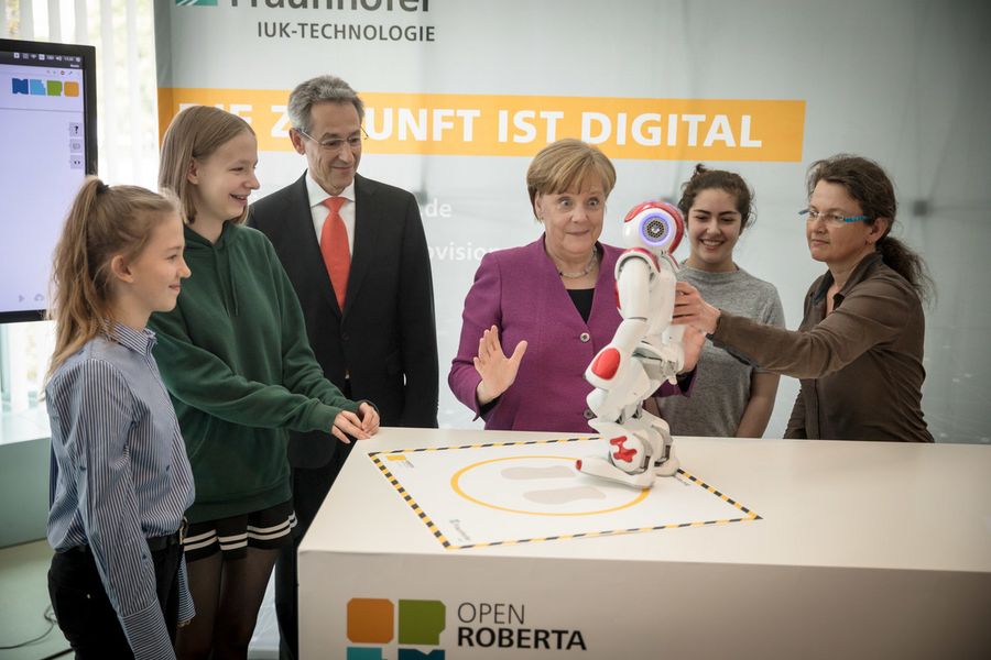 Foto: Eine Gruppe von sechs Personen steht um einen Tisch auf dem ein Roboter steht. Eine Person ist Bundeskanzlerin Angela Merkel, die erstaunt auf den Roboter schaut.