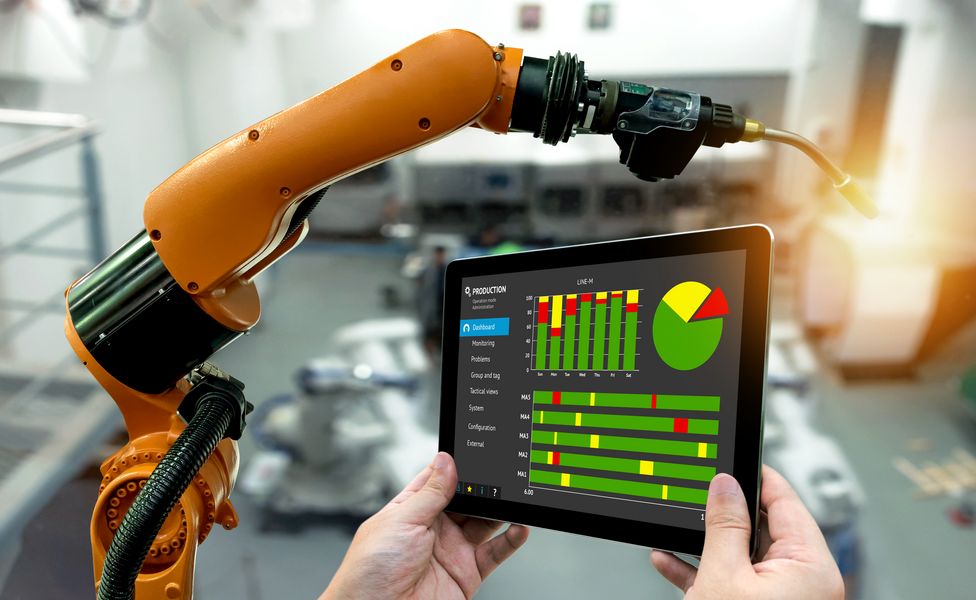 Tablet mit verschiedenen Diagrammen wird in Kamera gehalten, im Hintergrund ein Roboterarm in einer Fabrik