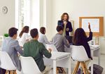 Klassenzimmer mit männlichem Leher im Vordergrund und zwei Bankreihen mit Schülerinnen und Schülern