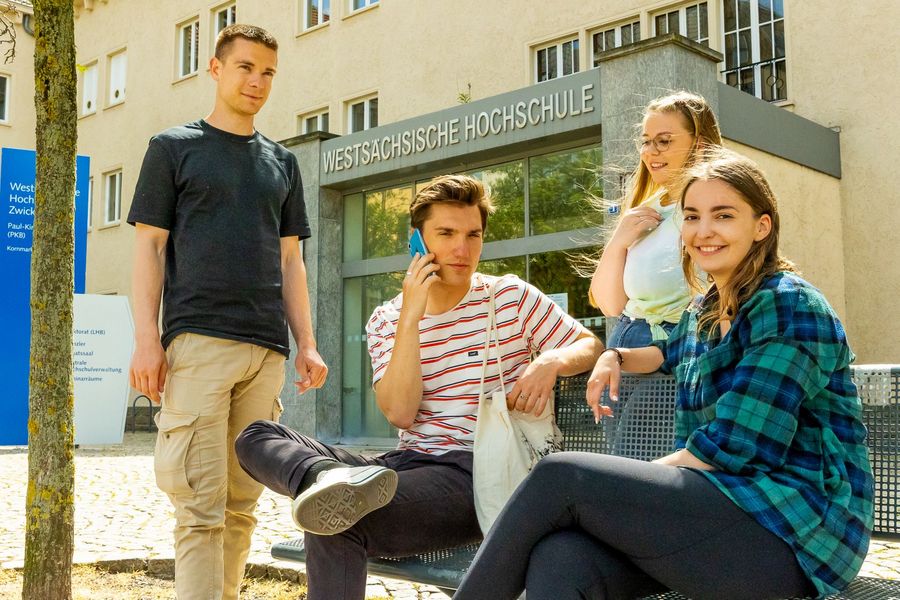 Foto: 4 junge Menschen stehen bzw. sitzen vor dem Portal der Westsächsischen Hochschule. 
