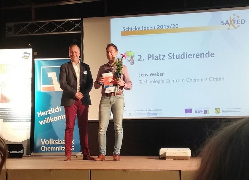 Foto: Zwei Personen auf einer Bühne stehend. Der Studierende Jens Weber erhält eine Urkunde und Blumen überreicht.