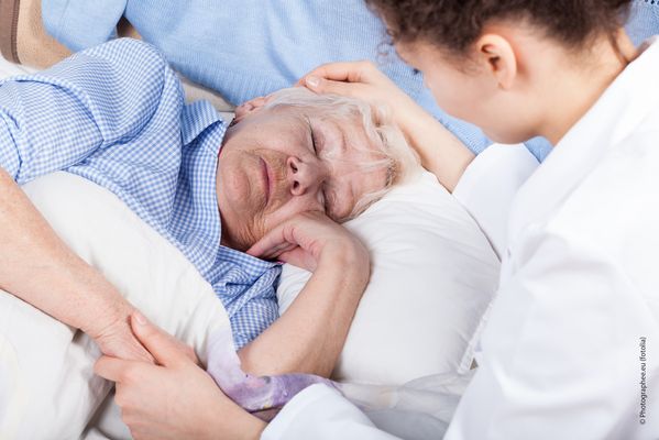 Symbolbild, Foto: Sterbebegleitung; Es ist eine ältere Frau zu sehen, die mit geschlossenen Augen in einem Bett liegt. Daneben steht eine Pflegerin, die der Frau übers Haar streicht. (Quelle: Fotolia)