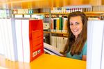 Foto: Eine Studierende lächelt durch eine Bücherregal Lücke.