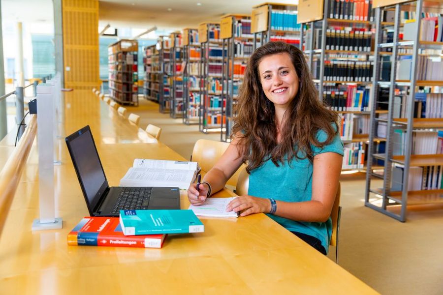 Foto: Eine Studentin sitzt lächelnd vor einem Laptop und Büchern in der Hochschulbilbliothek.