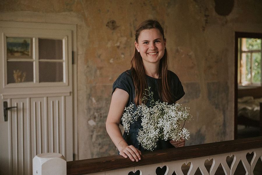Foto: Eine junge Frau steht mit einem Blumenstrauss lächelnd im Flur eines alten Hauses. 