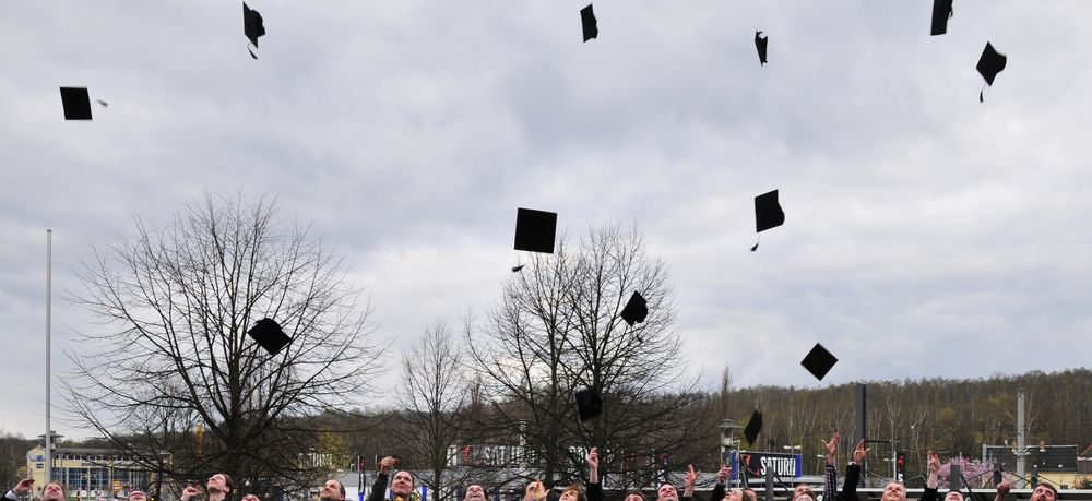 Foto: Examenshüte fliegen durch Luft. Eine Vielzahl Studierender stehen darunter, welche Sie zeitgleich in die Luft geworfen hatten.