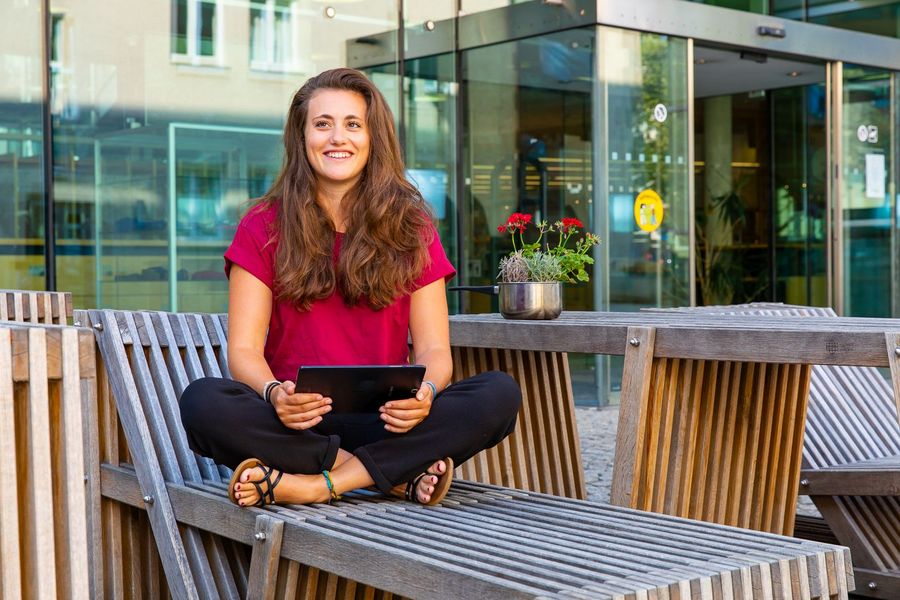 Foto: Eine Studierende sitzt draußen vor der Hochschulbibliothek und hält einen Laptop in der Hand.