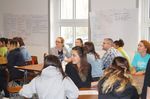 Foto: Studierende sitzen Gruppenweise in einem Lehrraum und hören einer Ansprache zu.