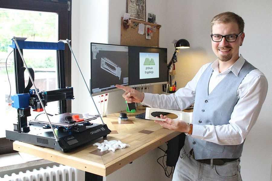 Foto: Ein junger Mann steht vor einem Schreibtisch. Auf dem sind ein 3D Drucker und zwei Monitore zu sehen. 