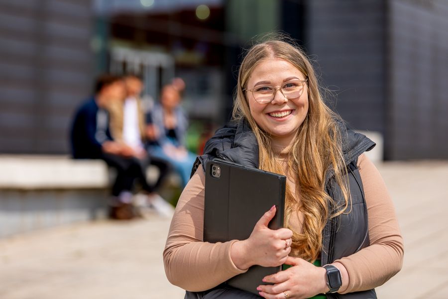 Eine Studentin auf dem Campus hält eine Mappe in der Hand und lächelt in die Kamera
