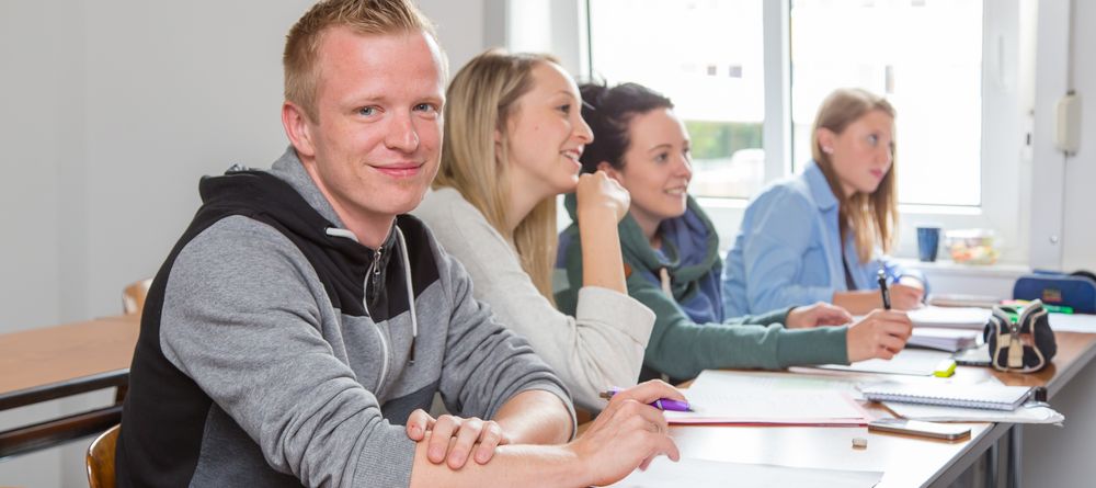 Foto: vier Studierende sitzen in einer Bankreihe eines Lehrraumes und hören einer Vorlesung zu.