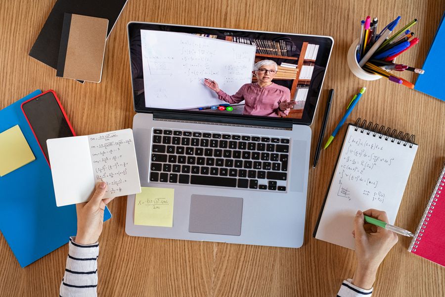 Foto: Laptop zeigt Online-Vorlesung Mathe, Zwei Hände, Stift und Notizblock, Stifte. 