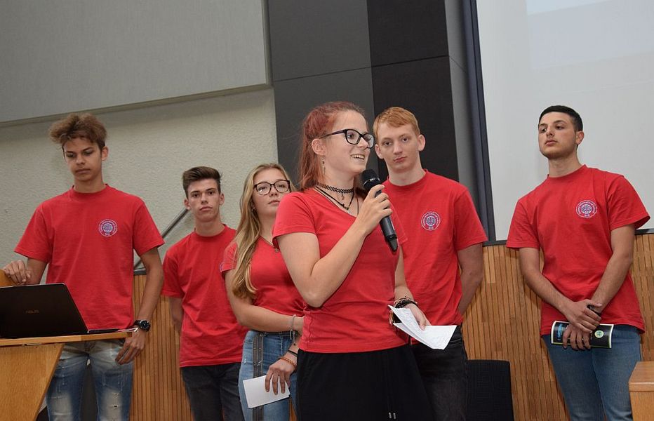Foto: Eine junge Studierende hält ein Mikrofon und spricht. Dahinter stehen 5 weitere Studierende der Gruppe.