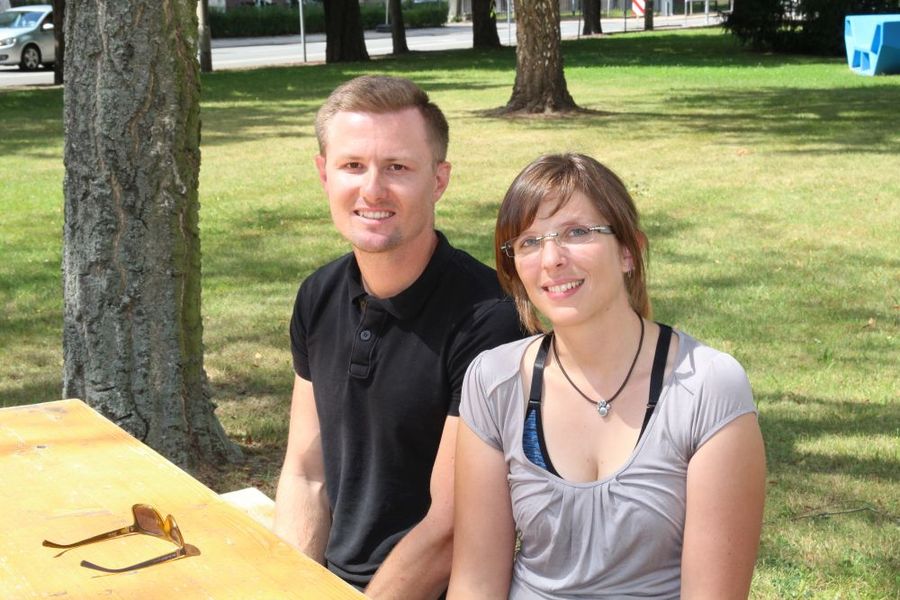 Foto: Ein junger Mann sitzt links, eine junge Frau rechts hinter einem Tisch. Im Hintergrund sind Wiese und Baumstämme.