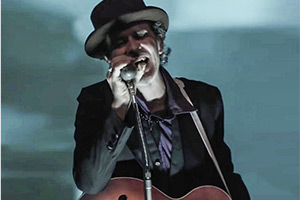 Foto: Ein Mann mit Gitarre um den Hals, singt in ein Mikrofon.