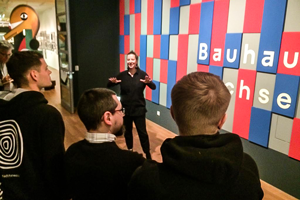 Foto: Beim Besuch im Bauhaus Sachsen stehen Studierende in einem Raum und hören einem Vortrag zu.