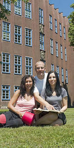 Foto: Drei Studierende sitzen auf einer Wiese vor dem Hochschulgebäude in Reichenbach und lächeln in die Kamera.
