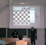 Foto 3: Zwei Studierende in einem Lehrraum, halten eine Präsentation. Auf dem Beamerbild an der Wand ist ein Schachbrett mit Figuren eingeblendet.