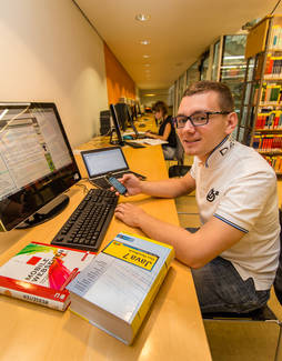 Foto: Ein Studierender sitzt an einem Rechercheplatz mit Computer in der Hauptbibliothek und lächelt in die Kamera.