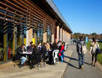 Foto 3: Studierende sitzen Tischen und Stühlen auf einer Terasse vor einem Gebäude.