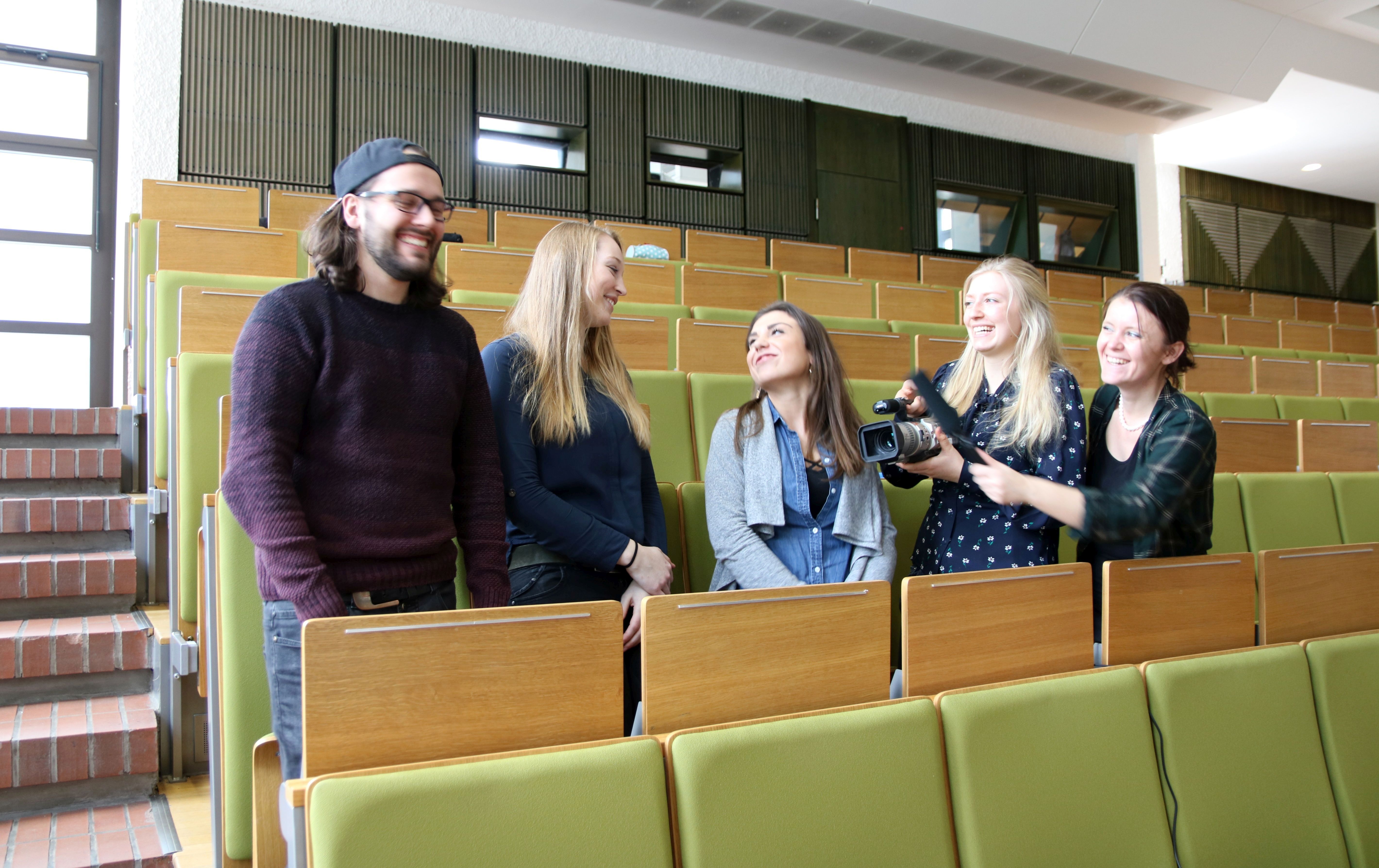 Foto: 5 Studierende stehen in einer Bankreihe des Hörsaal 2, lächeln und schauen sich gegenseitig an.