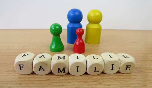 Symbolbild: Spielfiguren in verschiedenen Farben und Größen stehen nebeneinander. Davor liegen Würfel mit Buchstaben darauf. Die Würfel bilden das Wort Familie.
