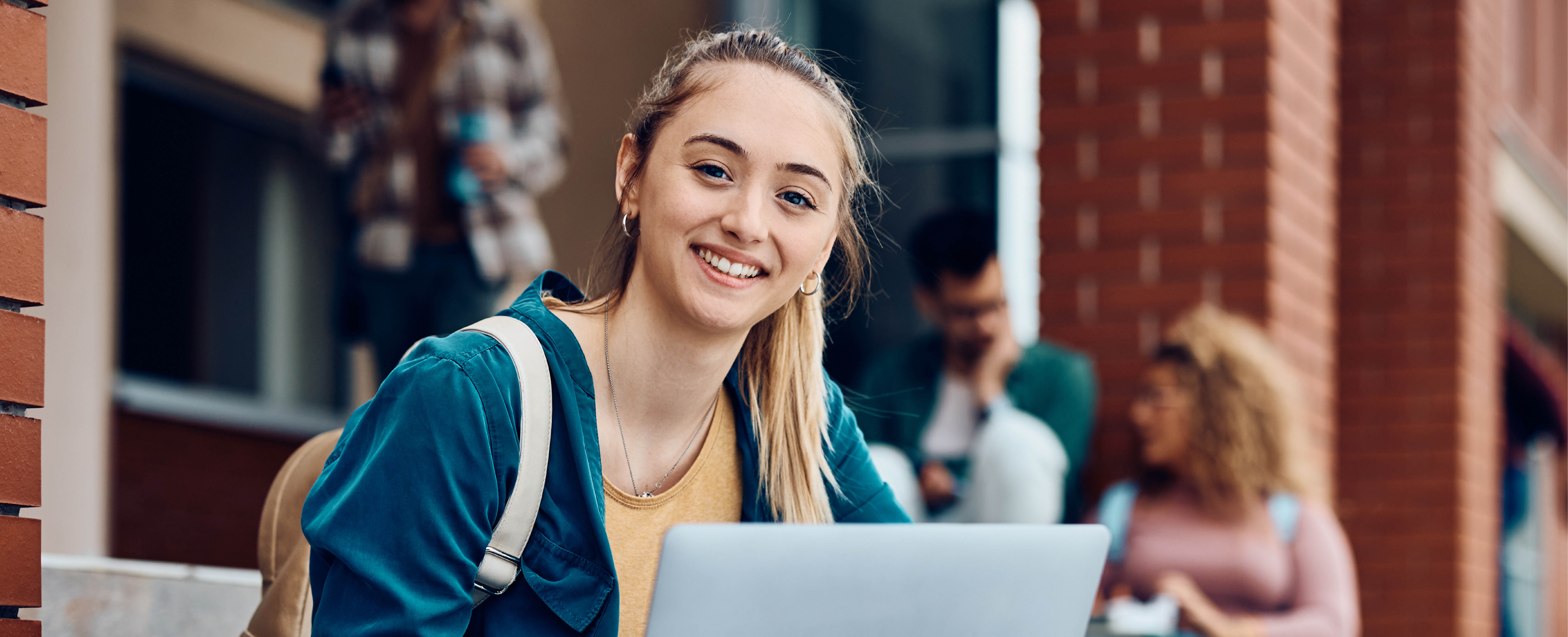 Studierende sitzt vor Ihren Notebook auf derm Campus und lächelt in die Kamera(Quelle: AdobeStock/Drazen) 