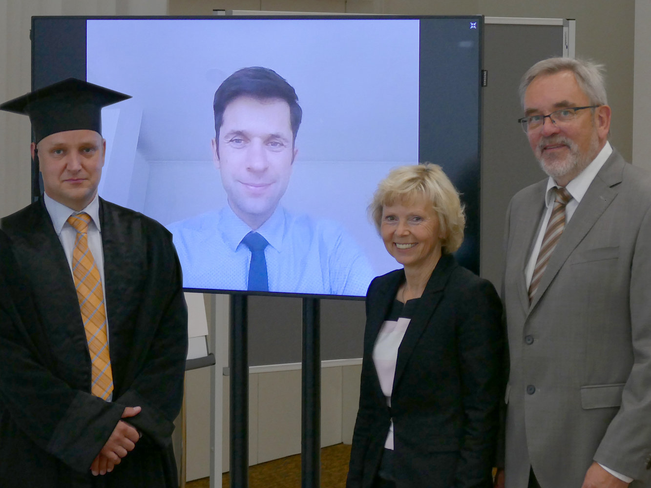 Der Doktorand links blickt mit seinen Betreuern in die Kamera, wobei ein Professor wegen Online-Teilnahme vom Monitor aus zu sehen ist.
