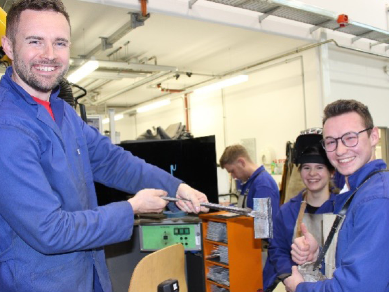 Männlicher Mitarbeiter und Studenten im blauen Kittel stehen vor einem Schweißstand im Labor und blicken lachend in die Kamera