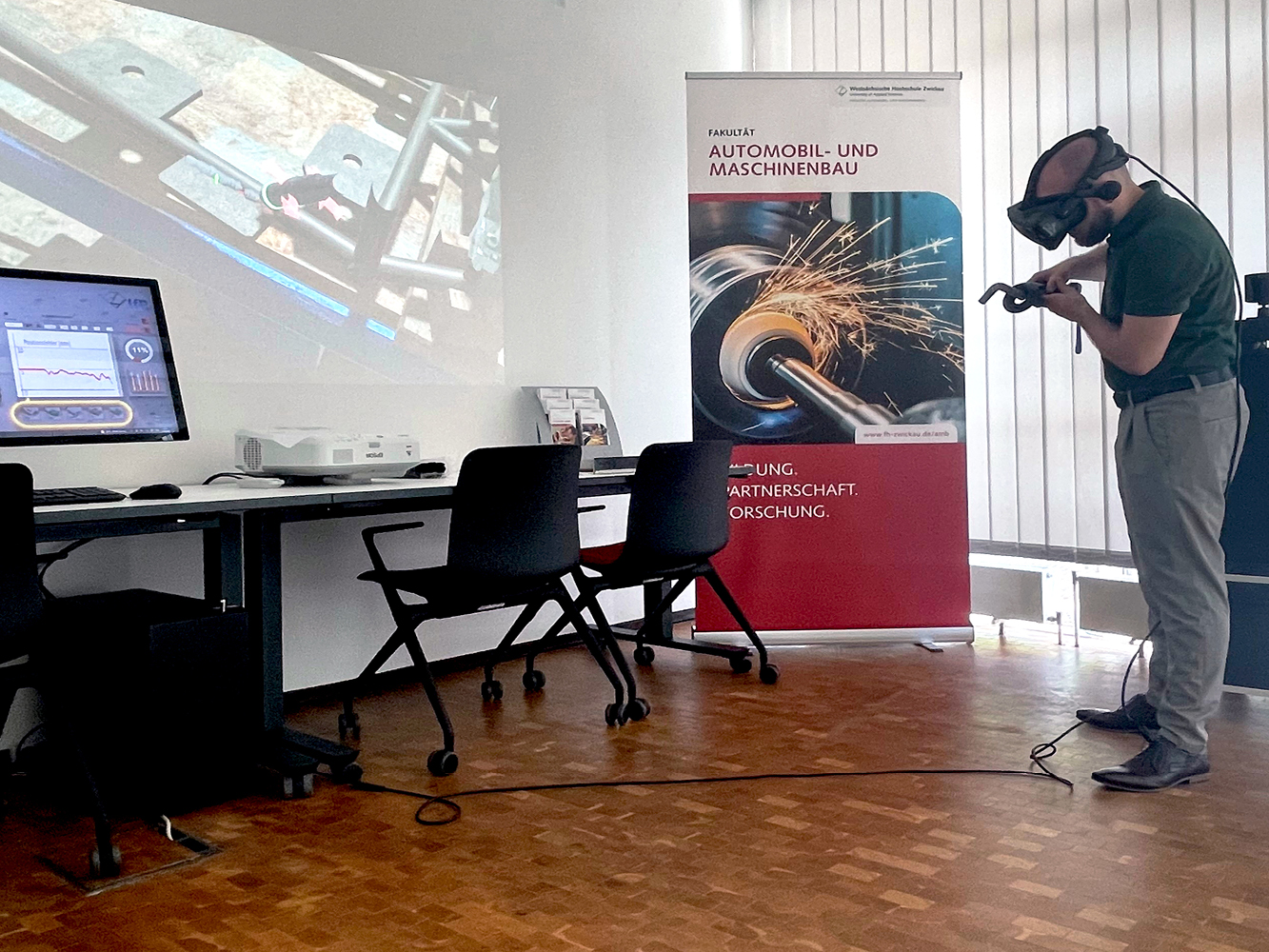 Mitarbeiter zeigt im Büro, wie virtuelles Schweißen mit VR-Brille funktioniert. Der digitale Vorgang ist auf der Wand links zu sehen. 