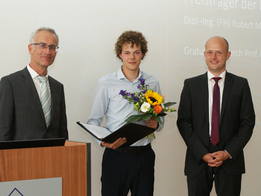 Absolvent steht mit Blumenstrauß und Urkunde in der Mitte, links der Dekan und rechts der betreuende Professor