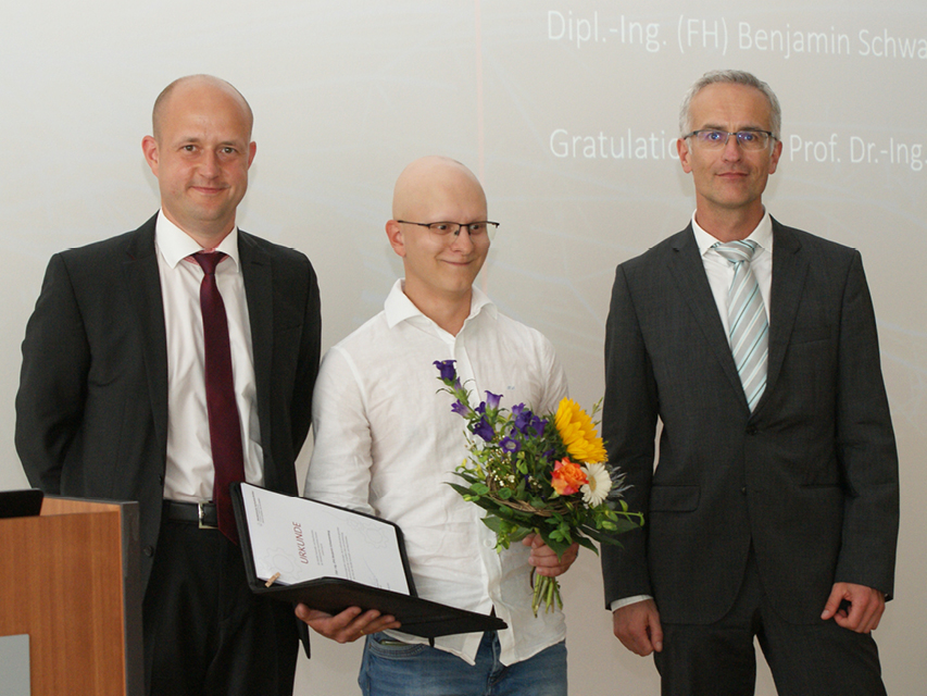 Absolvent steht mit Blumenstrauß und Urkunde in der Mitte, links der Dekan und rechts der betreuende Professor