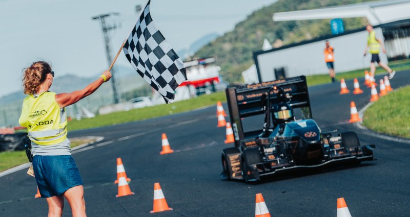 Der Rennwagen des WHZ Racingteams auf der Rennstrecke, links eine Frau mit weiß-schwarzer Fahne.