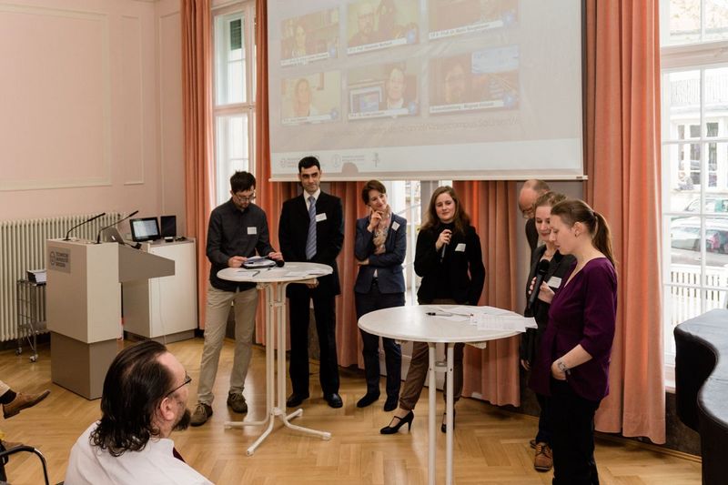 Foto: Zur Kickoff Veranstaltung der Umsetzungsphase des Projektes Videocampus Sachsen stehen sieben Teilnehmer um zwei Stehtische herum und führen einen offenen Dialog untereinander vor interessierten Publikum.