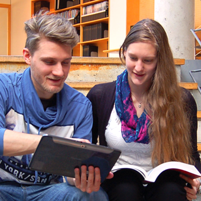 Foto: 2 Studierende mit einem Buch und einem Tablet unterhalten sich