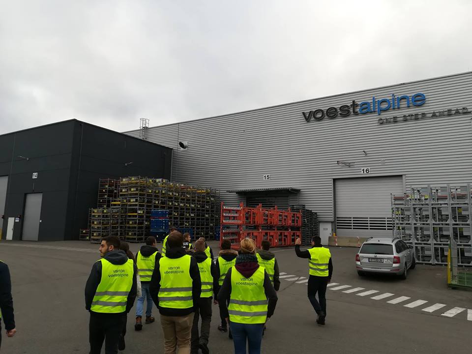 Foto: Eine Gruppe studierender mit Warnwesten laufen auf einem Vorplatz der Unternehmens Voestalpine zu einer Werkhalle.