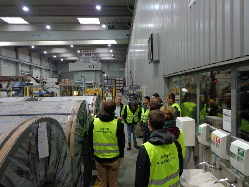 Foto: Eine Gruppe studierender steht in einer Werkhalle der Firma Voestalpine. Ein Mitarbeiter führt das Unternehmen vor.