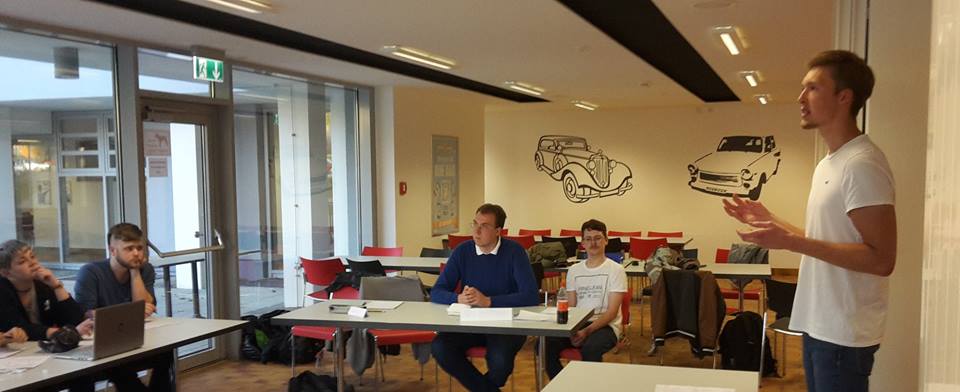 Foto: Studierende sitzen an einer U-förmigen Tischanordnung in der Mensa Scheffelberg. Ein Studierender hält stehend eine Rede.