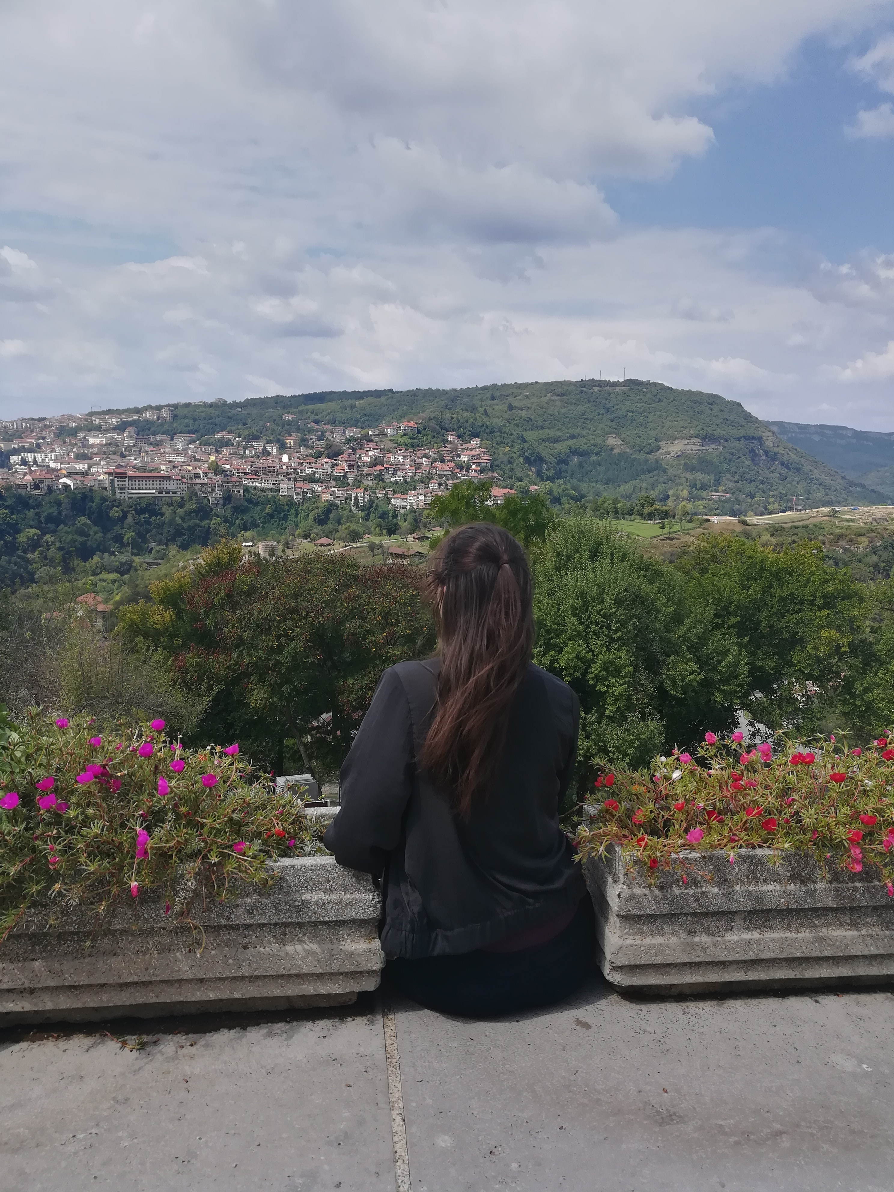 Foto: Ein Frau sitzt mit dem Rücken zur Kamera am Rand eines Hügelplattform und schaut über die hügelige Landschaft in der Ferne.
