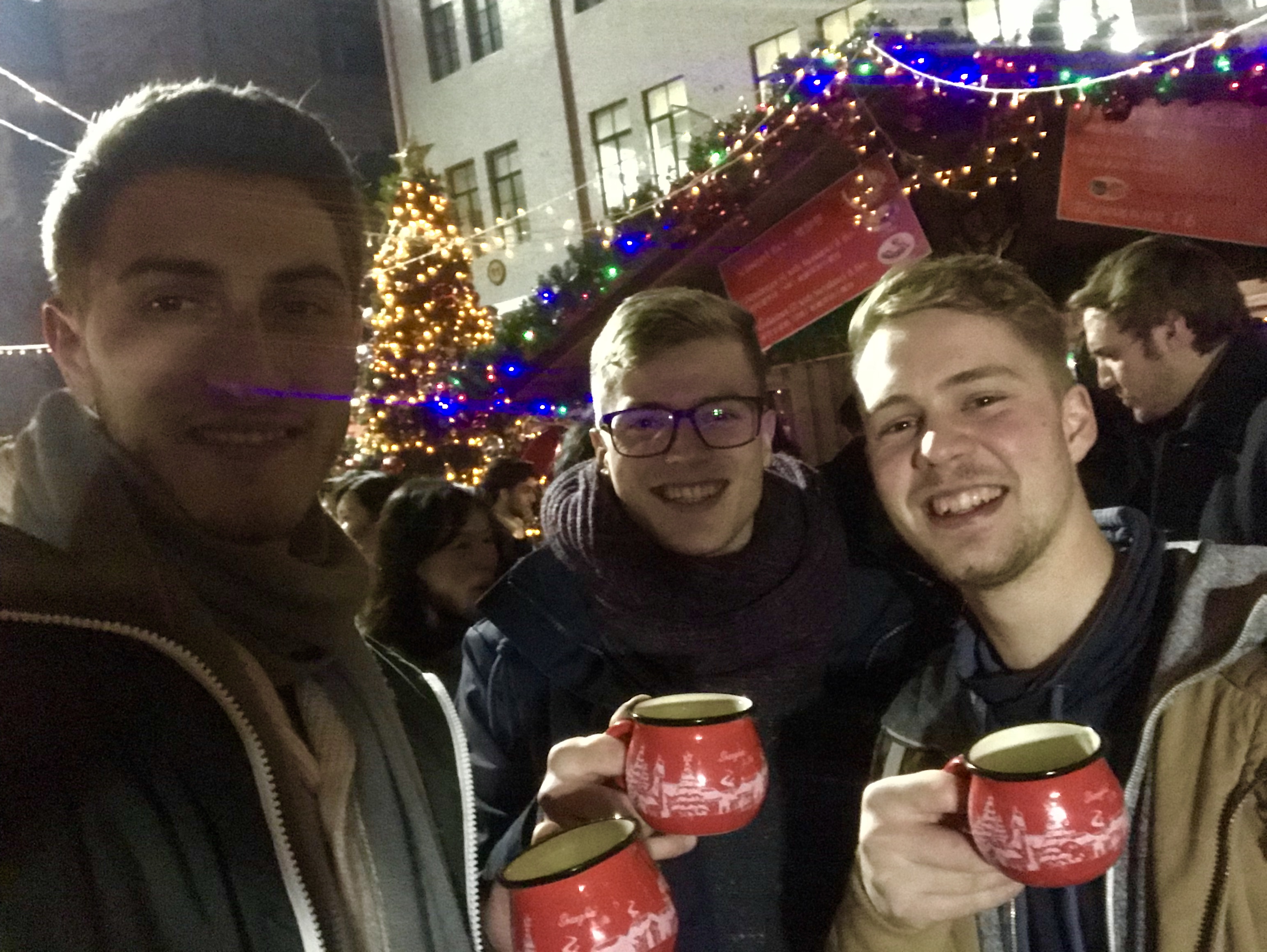 Foto: Drei Studierende mit Glühweintassen in der Hand tätigen ein Selfie Foto auf dem Weihnachtsmarkt.