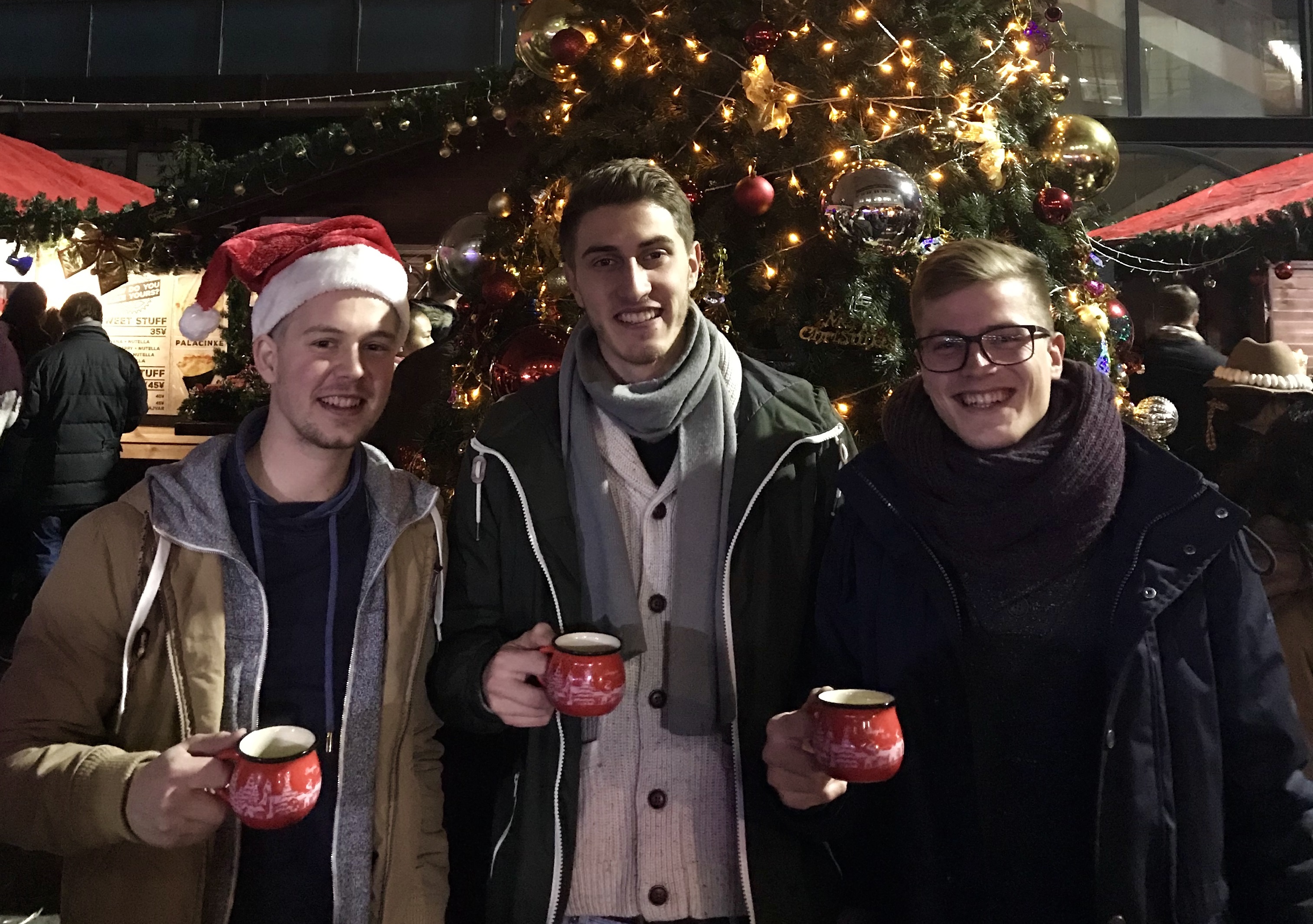 Foto: Drei Studierende mit Glühweintassen in der Hand tätigen ein Gruppenbild auf dem Weihnachtsmarkt.