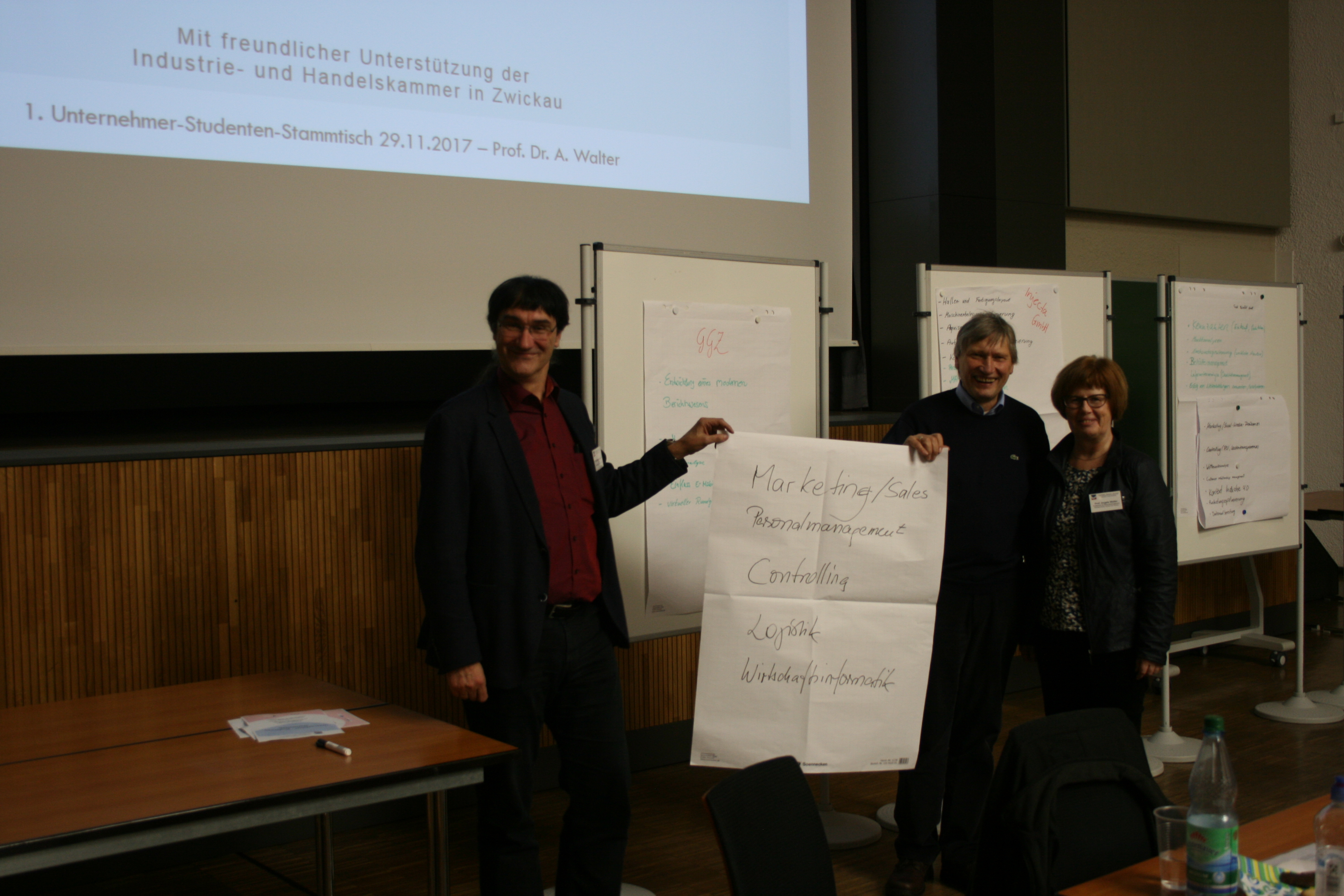 Foto: 1. Unternehmer-Studenten-Stammtisch. Drei Professoren stehen vor einer Reihe Flipcharts und halten ein beschriftetes Plakat in die Kamera.